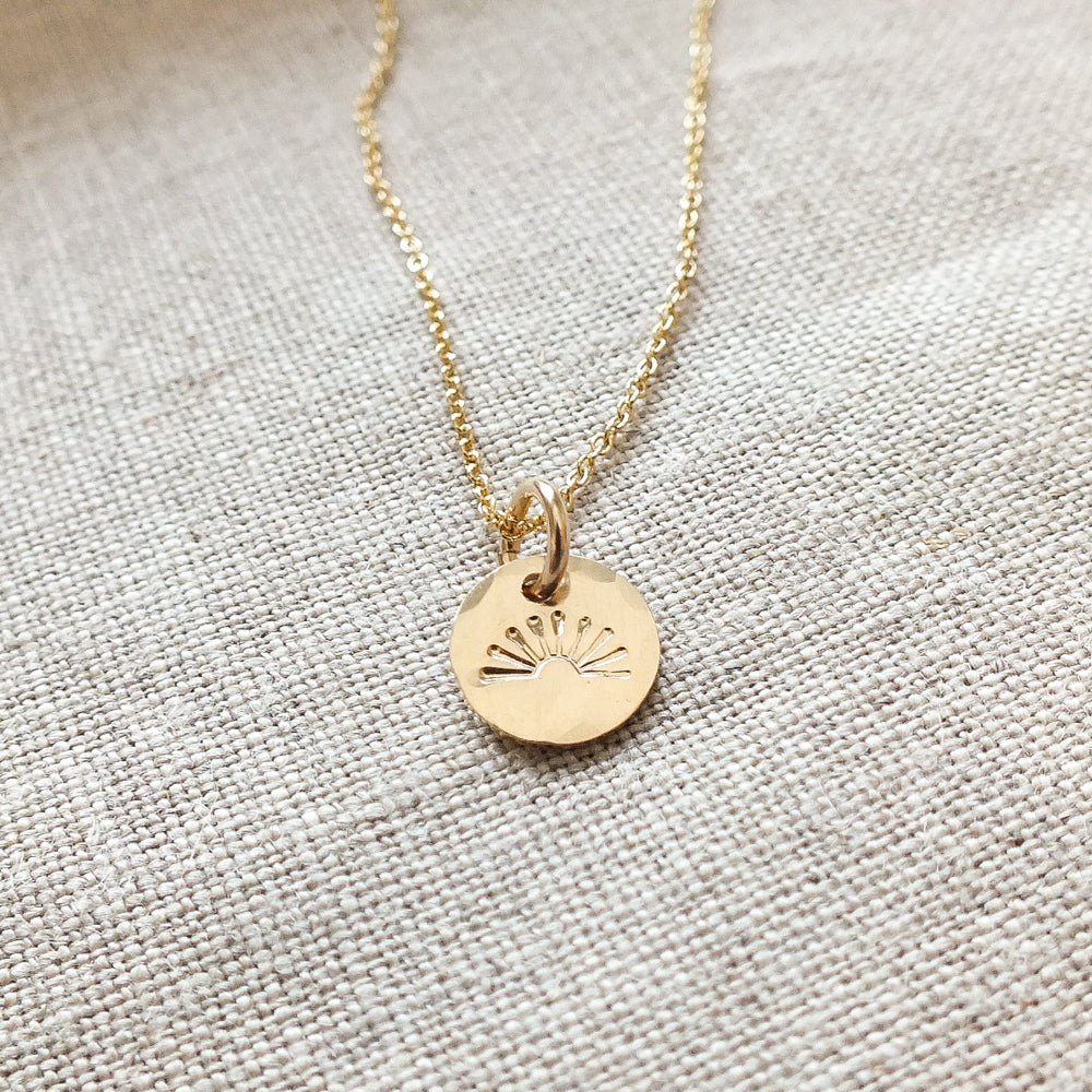 You Are My Sunshine Necklace by Becoming Jewelry, showcasing a radiant sun design on a round pendant and displayed on a textured fabric background. This bestselling necklace makes for the perfect gift.