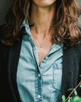 A person with long, wavy hair wears a denim shirt under a black cardigan, with the bestselling You Are My Sunshine Necklace by Becoming Jewelry—a small circular pendant that gleams like a radiant sun—visible.