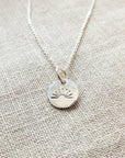 The You Are My Sunshine Necklace by Becoming Jewelry, a bestselling piece, showcases a silver round pendant with an engraved radiant sun design, elegantly displayed on a light fabric background.