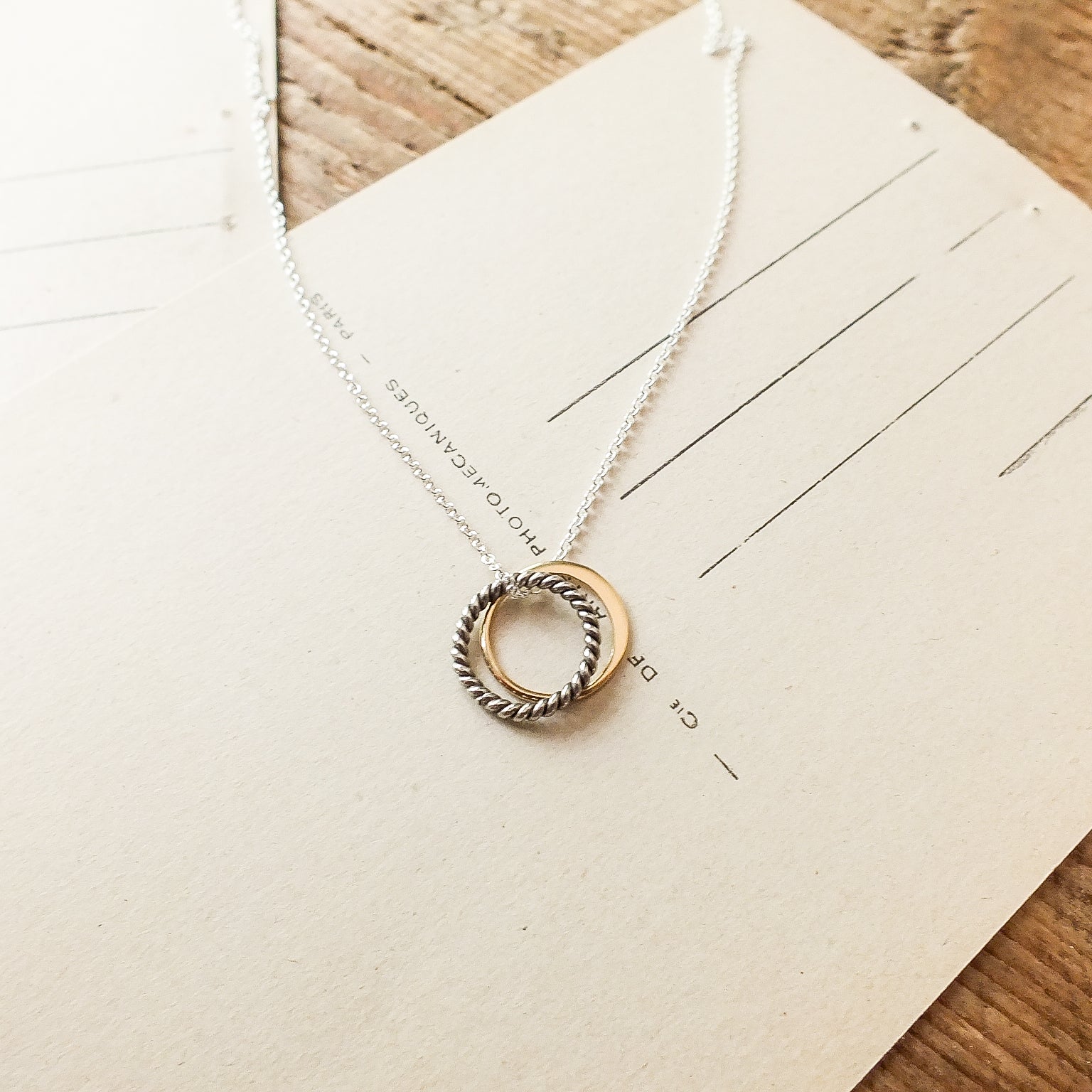 The True Friends Necklace by Becoming Jewelry, featuring two mixed metal textured circles—one gold and one silver—adorns an off-white envelope set on a wooden surface.