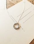 The True Friends Necklace by Becoming Jewelry, featuring two mixed metal textured circles—one gold and one silver—adorns an off-white envelope set on a wooden surface.