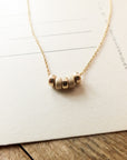 A close-up of the **My Wish For You Necklace** by **Becoming Jewelry**, a delicate gold piece featuring five small cylindrical beads, elegantly displayed on a paper background—an ideal choice for a thoughtful gift.