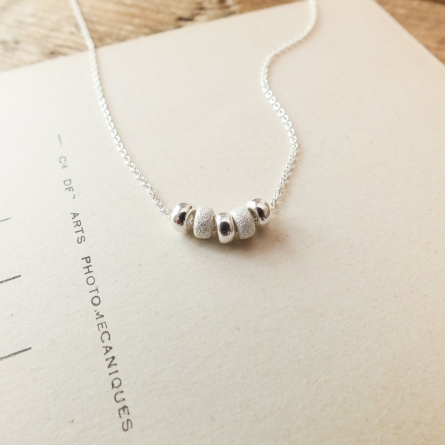 Becoming Jewelry&#39;s My Wish For You Necklace, a sterling silver comfort charm necklace with three beads, is featured on a wooden surface against a paper background.