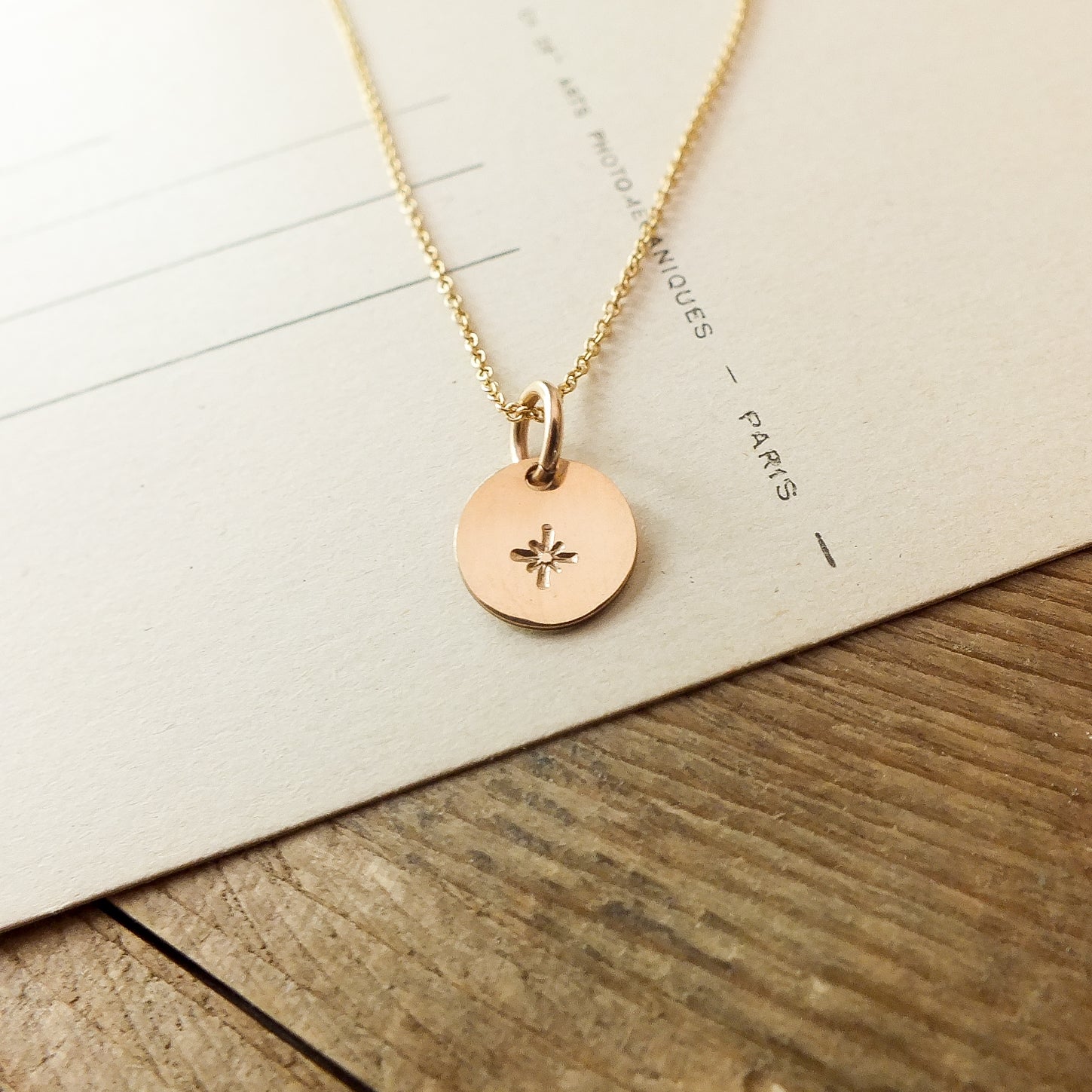 The Light Within Necklace by Becoming Jewelry features a gold pendant with a dainty starburst symbol on a circular charm, set against a paper background with visible text. The chain of the necklace is thin and delicate, adding to its elegance.