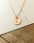 The Light Within Necklace by Becoming Jewelry features a gold pendant with a dainty starburst symbol on a circular charm, set against a paper background with visible text. The chain of the necklace is thin and delicate, adding to its elegance.