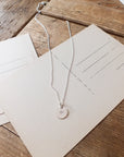 The Light Within Necklace by Becoming Jewelry, a silver pendant adorned with a tiny starburst, lay on top of a blank postcard on a wooden surface.