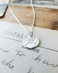 The Compass Necklace by Becoming Jewelry, featuring a compass charm pendant, displayed over postcards and handwritten text, offers travel inspiration.