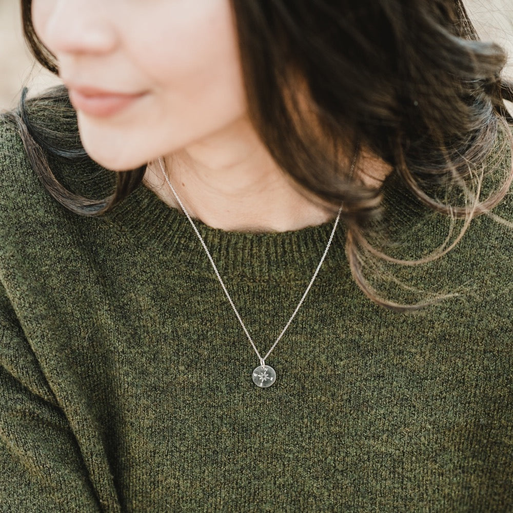 A person is shown from the shoulders up, facing slightly left, wearing a green sweater and Becoming Jewelry's Compass Necklace with a round pendant. The background is blurred, highlighting the charm on the pendant and evoking a sense of travel inspiration.