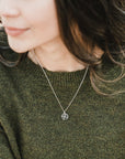A person is shown from the shoulders up, facing slightly left, wearing a green sweater and Becoming Jewelry's Compass Necklace with a round pendant. The background is blurred, highlighting the charm on the pendant and evoking a sense of travel inspiration.