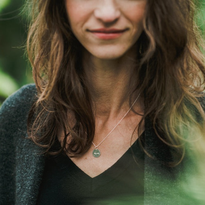 A woman with wavy brown hair and a subtle smile is wearing the Arrow Necklace from Becoming Jewelry. She is dressed in a dark top and a gray cardigan, standing outdoors with blurred greenery in the background, embodying quiet motivation and encouragement.