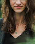 A woman with wavy brown hair and a subtle smile is wearing the Arrow Necklace from Becoming Jewelry. She is dressed in a dark top and a gray cardigan, standing outdoors with blurred greenery in the background, embodying quiet motivation and encouragement.