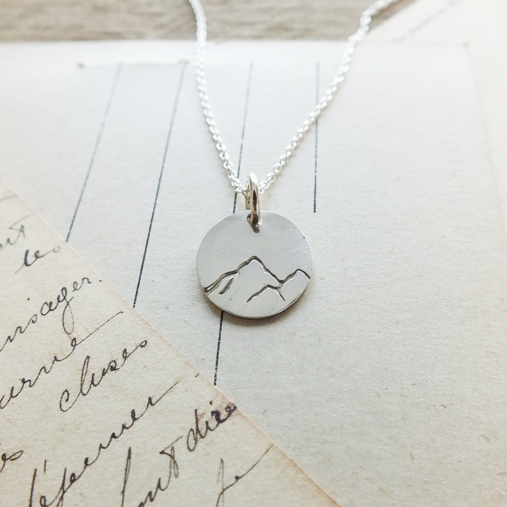 Sterling silver Mountains Are Calling necklace charm pendant on a necklace chain placed on handwritten paper by Becoming Jewelry.