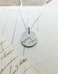 The "Mountains Are Calling Necklace" by Becoming Jewelry, a silver necklace with a small round pendant featuring an etched mountain design, is elegantly displayed on a piece of handwritten paper, evoking a sense of nature and adventure.