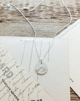 The "Mountains Are Calling Necklace" by Becoming Jewelry, featuring a small silver round pendant engraved with mountain peaks, is displayed on a wooden surface alongside vintage postcards, evoking a sense of adventure and the beauty of nature.