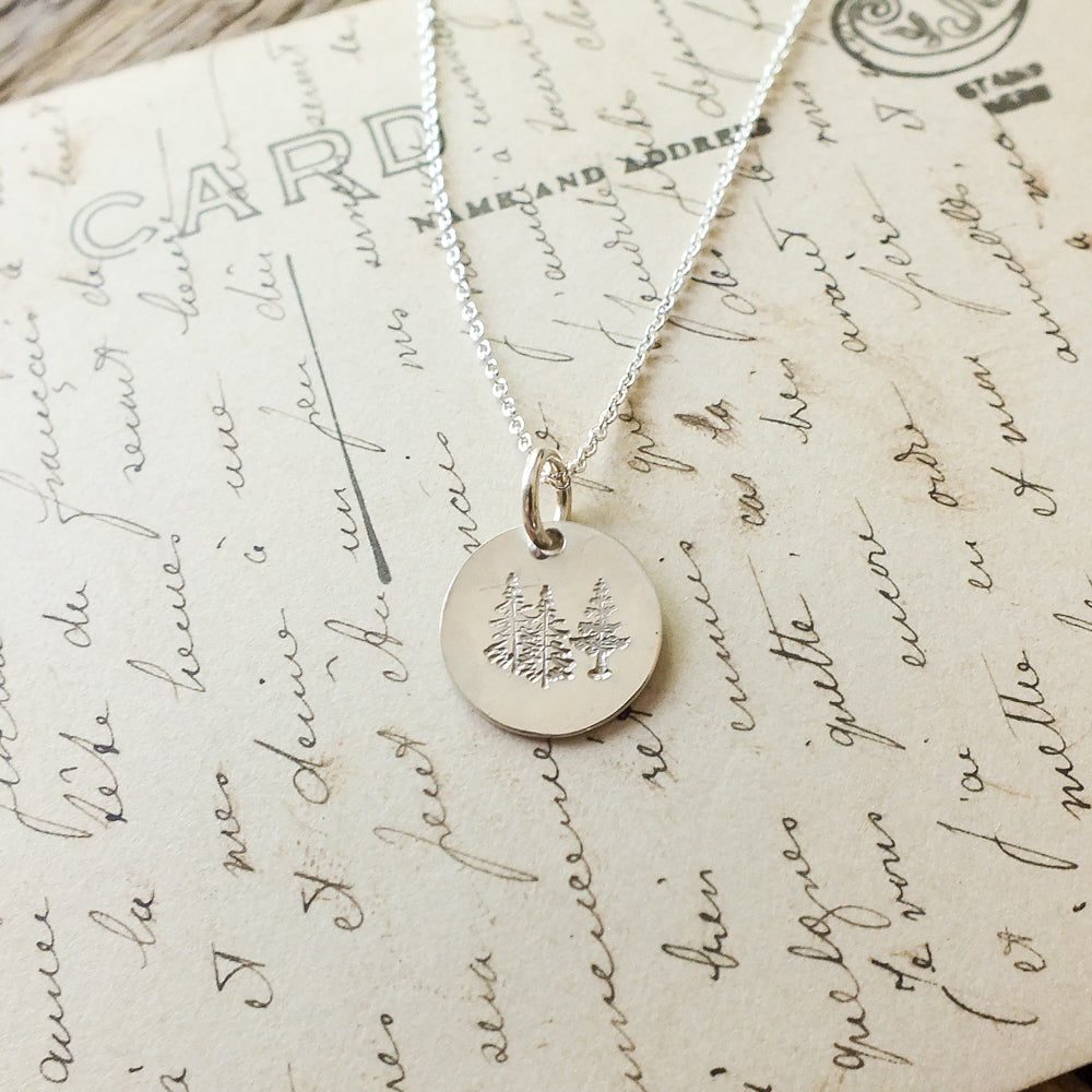 The Trees Necklace from Becoming Jewelry, a silver necklace adorned with a circular pendant featuring an engraved tree design, is elegantly placed on a vintage handwritten letter, capturing the essence of nature's restoration.