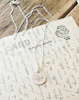 The "Trees Necklace" by Becoming Jewelry, featuring a nature-inspired tree pendant in silver, rests on handwritten postcards on a wooden surface.