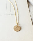 The River Necklace by Becoming Jewelry, featuring a gold round pendant engraved with an abstract design and presented on a piece of beige paper bearing fine markings, embodies the indomitable spirit of remarkable women.