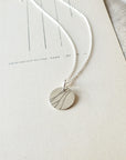 A silver River Necklace by Becoming Jewelry, featuring a round pendant with minimalist line designs, rests on an off-white textured surface with faint lines, symbolizing the indomitable spirit of remarkable women.
