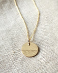 The Sea Necklace by Becoming Jewelry features a gold round pendant engraved with a minimalistic wave pattern, reminiscent of the ocean, displayed on a textured fabric background.