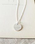 Close-up of The Sea Necklace by Becoming Jewelry, featuring a round pendant with an ocean wave pattern, placed on a beige textured background.