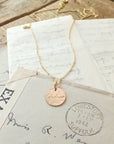 The Badass Necklace by Becoming Jewelry, a gold necklace with a round pendant engraved in an antique handwriting font with the name "Isabella," rests on top of vintage letters, one of which is postmarked from Lowestoft, Suffolk, dated 1942. This piece exudes empowerment as it connects a badass sense of history and legacy.
