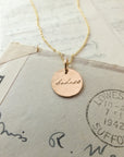 The "Badass Necklace" by Becoming Jewelry features a gold disc pendant engraved with the word "badass" in an antique handwriting font. Placed on top of an old stamped letter dated September 7, 1942, sent to Lowestoft, Suffolk, this unique piece radiates a sense of empowerment and timeless elegance.