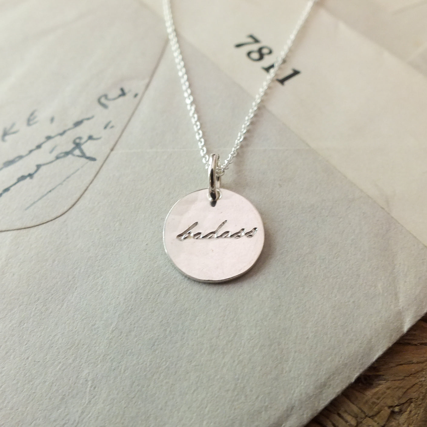 A Becoming Jewelry Badass Necklace with the word &quot;badass&quot; engraved, resting on an envelope.