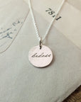 The Badass Necklace from Becoming Jewelry, featuring a round pendant inscribed with "badass" in an antique handwriting font, rests on a tan envelope, radiating a sense of empowerment.