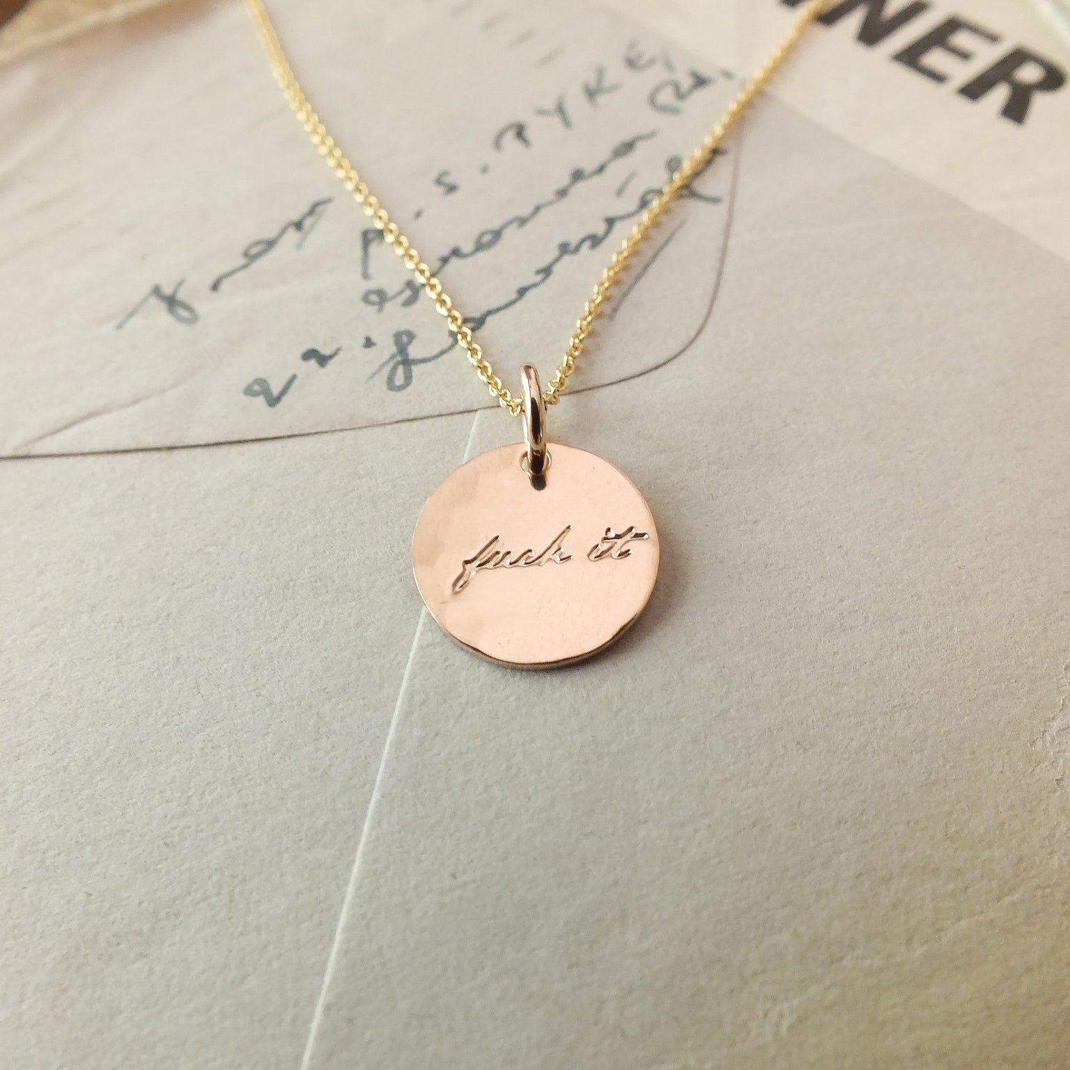 A gold-filled Fuck It Necklace by Becoming Jewelry with a circular pendant inscribed with the phrase &quot;just do it&quot; resting on a paper surface with handwritten text.