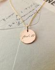 A gold-filled Fuck It Necklace by Becoming Jewelry with a circular pendant inscribed with the phrase "just do it" resting on a paper surface with handwritten text.