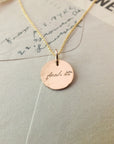 A round gold pendant necklace from Becoming Jewelry’s "Fuck It Necklace" collection, engraved with the cursive phrase "fuck it," is displayed atop an envelope adorned with antique handwriting font.