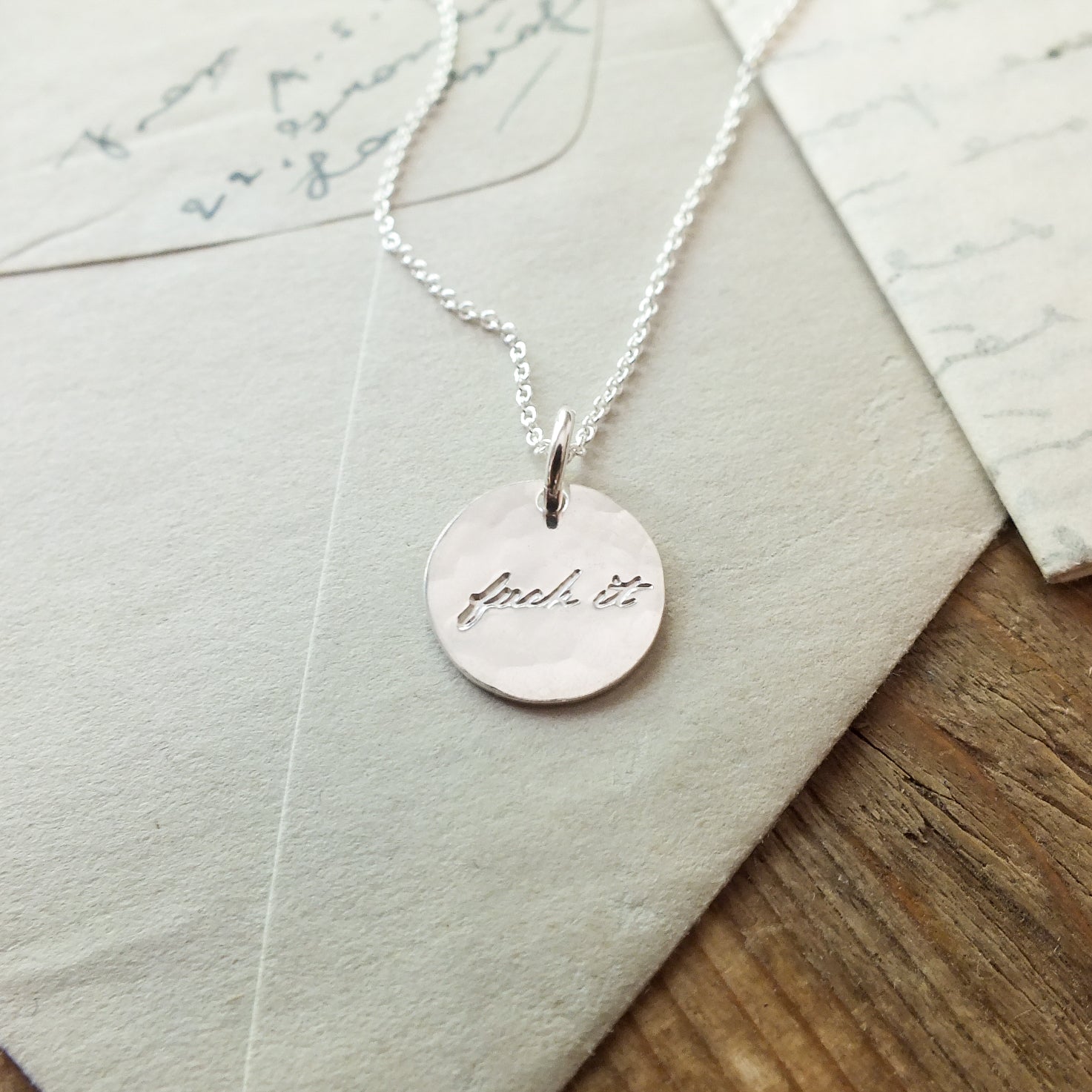 Becoming Jewelry&#39;s Fuck It Necklace with handwritten inscription on wooden background with envelopes.