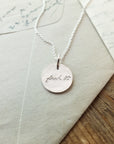 A silver necklace called the Fuck It Necklace from Becoming Jewelry, featuring a small round charm engraved in an antique handwriting font with the inspirational words "fuck it," rests on cream-colored envelopes and notecards.