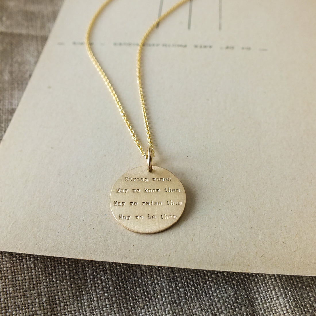 Becoming Jewelry&#39;s Strong Women Necklace is a sterling silver charm necklace with an engraved inspirational quote for strong women on a beige background.