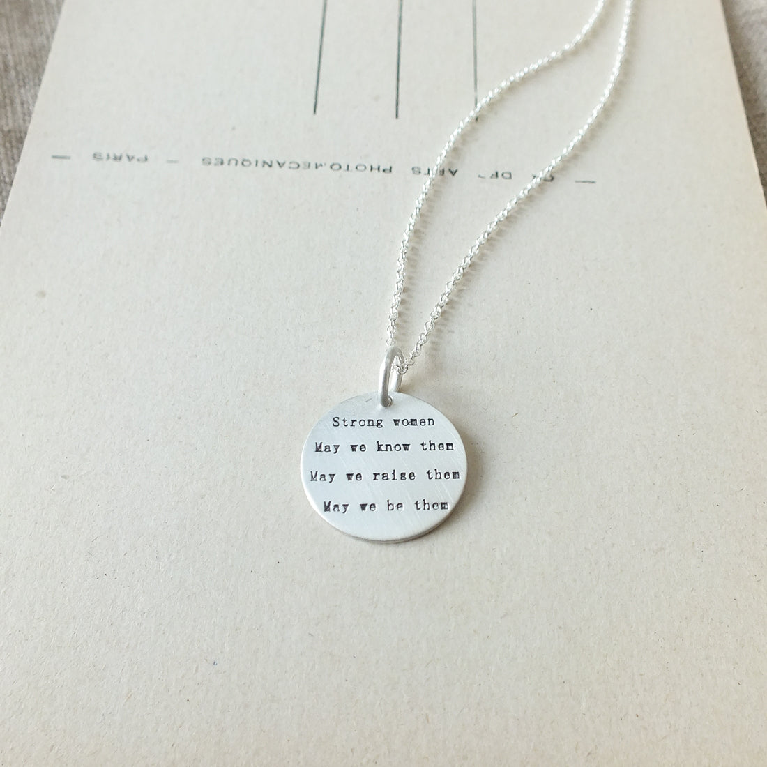 Becoming Jewelry&#39;s Strong Women Necklace with a charm inscribed with a message about strong women displayed on a piece of paper.