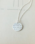 Presenting the Strong Women Necklace by Becoming Jewelry: a round silver pendant on a chain, engraved with the empowering message, "Strong Women. May we know them. May we raise them. May we be them." Ideal for daily wear or as a thoughtful gift, this elegant piece is beautifully presented on a sheet of paper.
