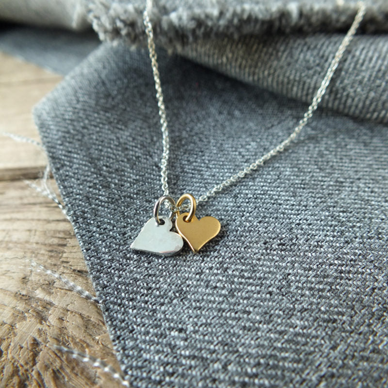 Close-up of the Tiny Hearts Necklace by Becoming Jewelry, showcasing mixed metal hearts—one silver and one gold—beautifully placed on grey fabric. The background features more grey fabric and a wooden surface, making it truly the perfect gift.