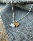 Close-up of the Tiny Hearts Necklace by Becoming Jewelry, showcasing mixed metal hearts—one silver and one gold—beautifully placed on grey fabric. The background features more grey fabric and a wooden surface, making it truly the perfect gift.