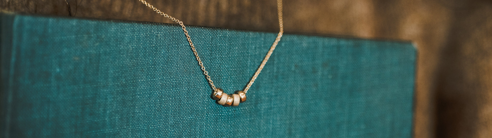 A delicate gold necklace with a pearl pendant hangs against a textured teal book cover.