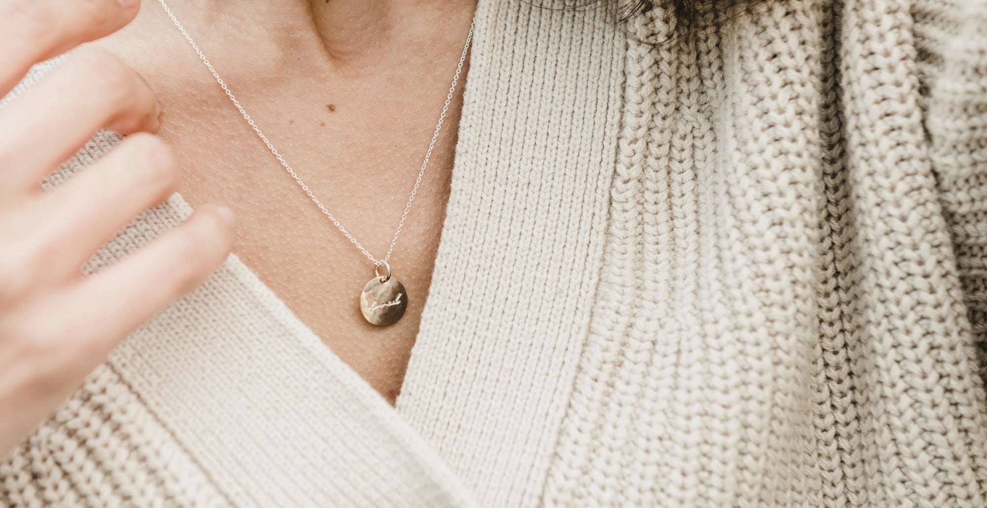 Close-up of a person wearing a beige sweater and a silver necklace with a pendant, with a hand slightly visible.