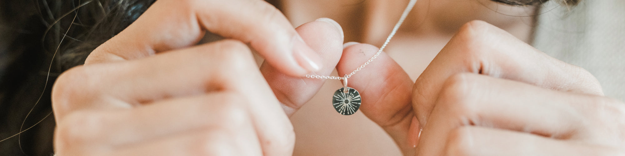 Close-up of a person wearing a beige sweater and a silver necklace with a pendant, with a hand slightly visible.