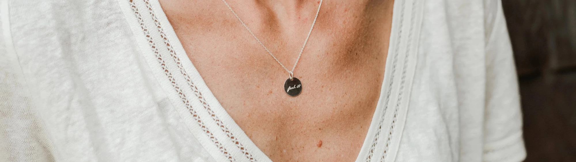 Close-up of a person wearing a white v-neck shirt with a silver necklace featuring a round pendant that says "mama.