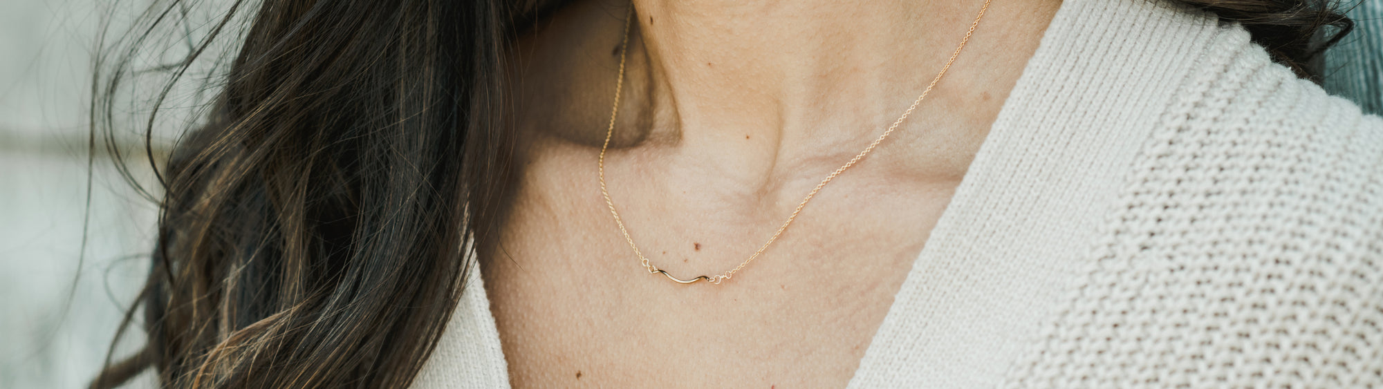 Close-up of a woman's neck wearing a delicate gold necklace with a small pendant, contrasted against her white sweater and dark hair.