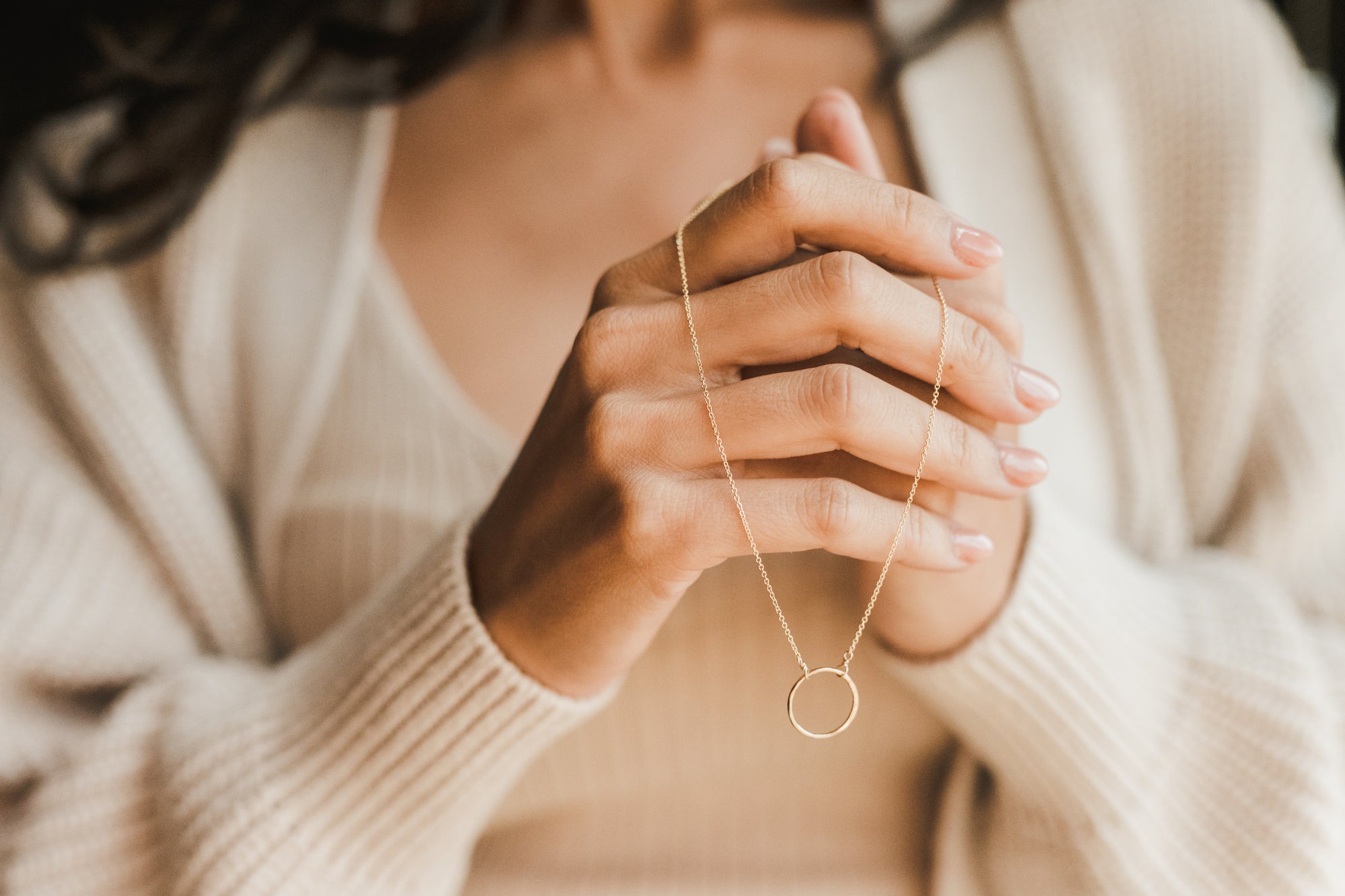 A woman in a cream sweater clasping a delicate pendant necklace.