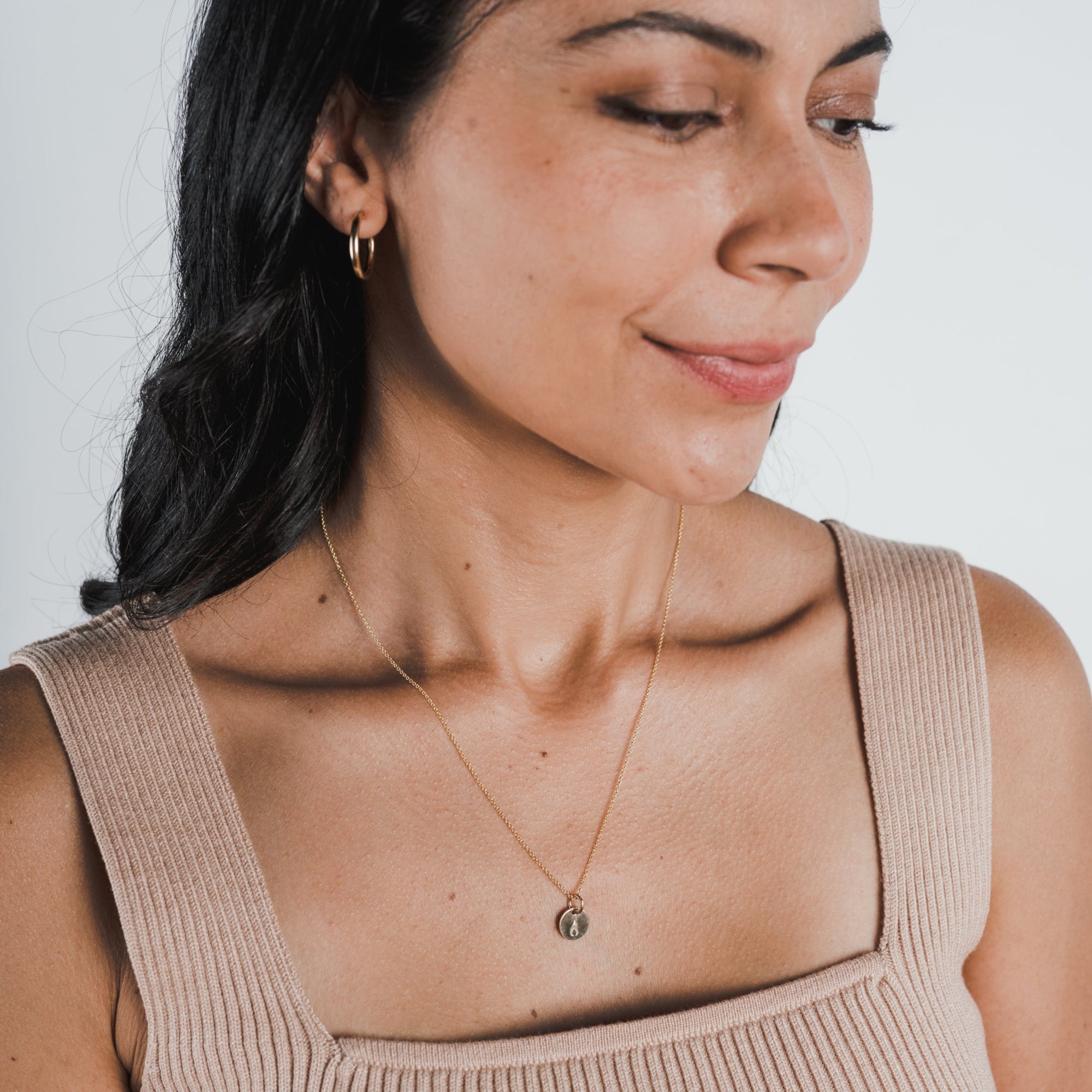 A woman with long dark hair is wearing a sleeveless beige top, small hoop earrings, and a delicate gold necklace with a small pendant. She is looking down and smiling slightly, perhaps reflecting on her motivation for the future. An On Fire Necklace from Becoming Jewelry makes an ideal gift for graduates like her.