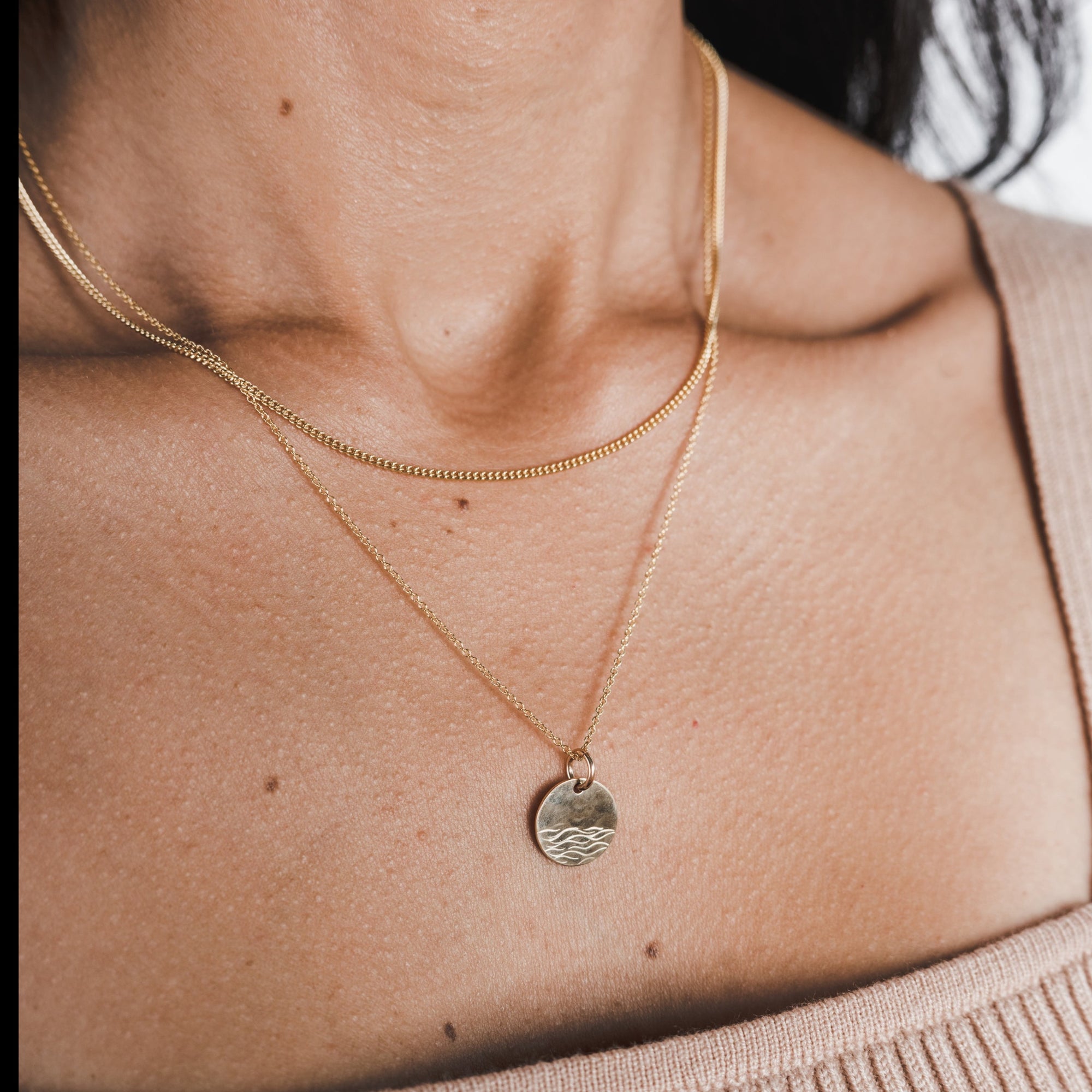Close-up of a person wearing layered necklaces, including the classic Curb Chain Necklace from Becoming Jewelry and a gold necklace with a round pendant featuring a wave design. The person is dressed in a light beige top.