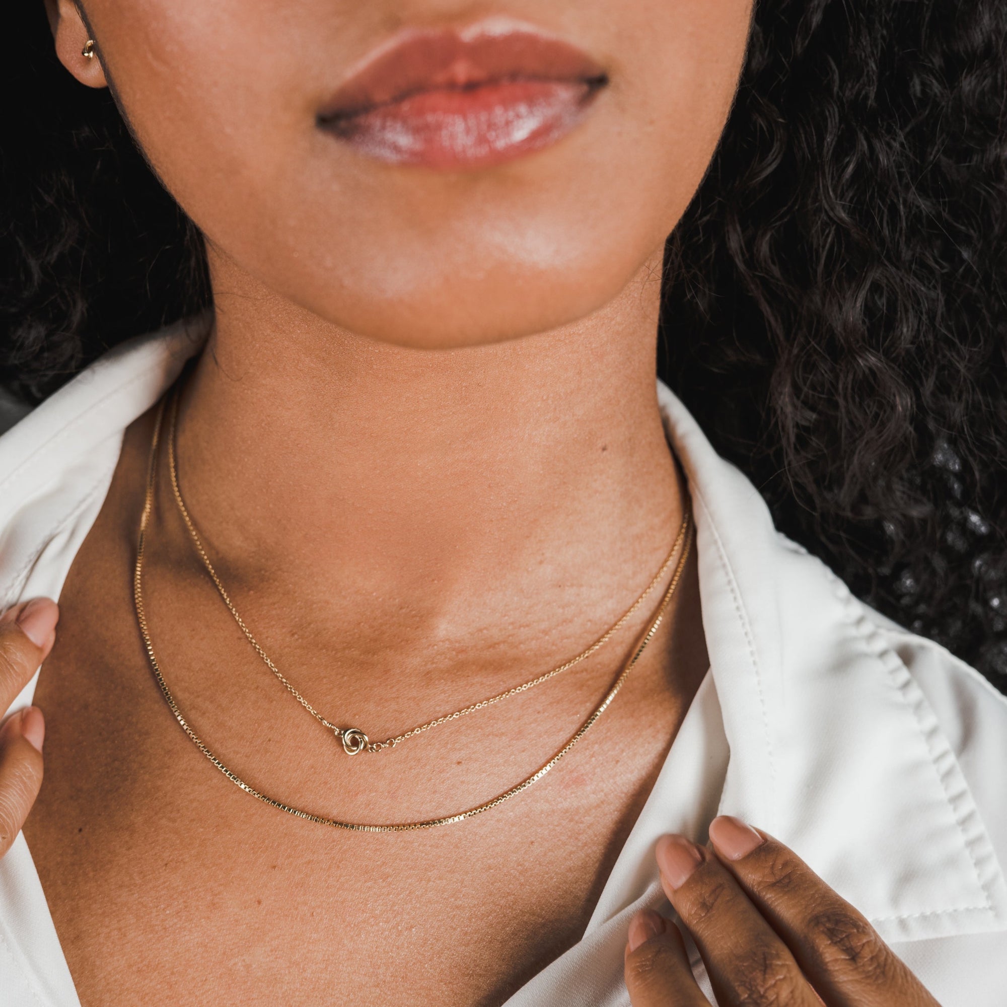 Close-up of a person wearing two gold necklaces and a white shirt, exuding a classic look. The neck and part of the face are visible, with hands gently holding the lightweight Box Chain Necklace by Becoming Jewelry.