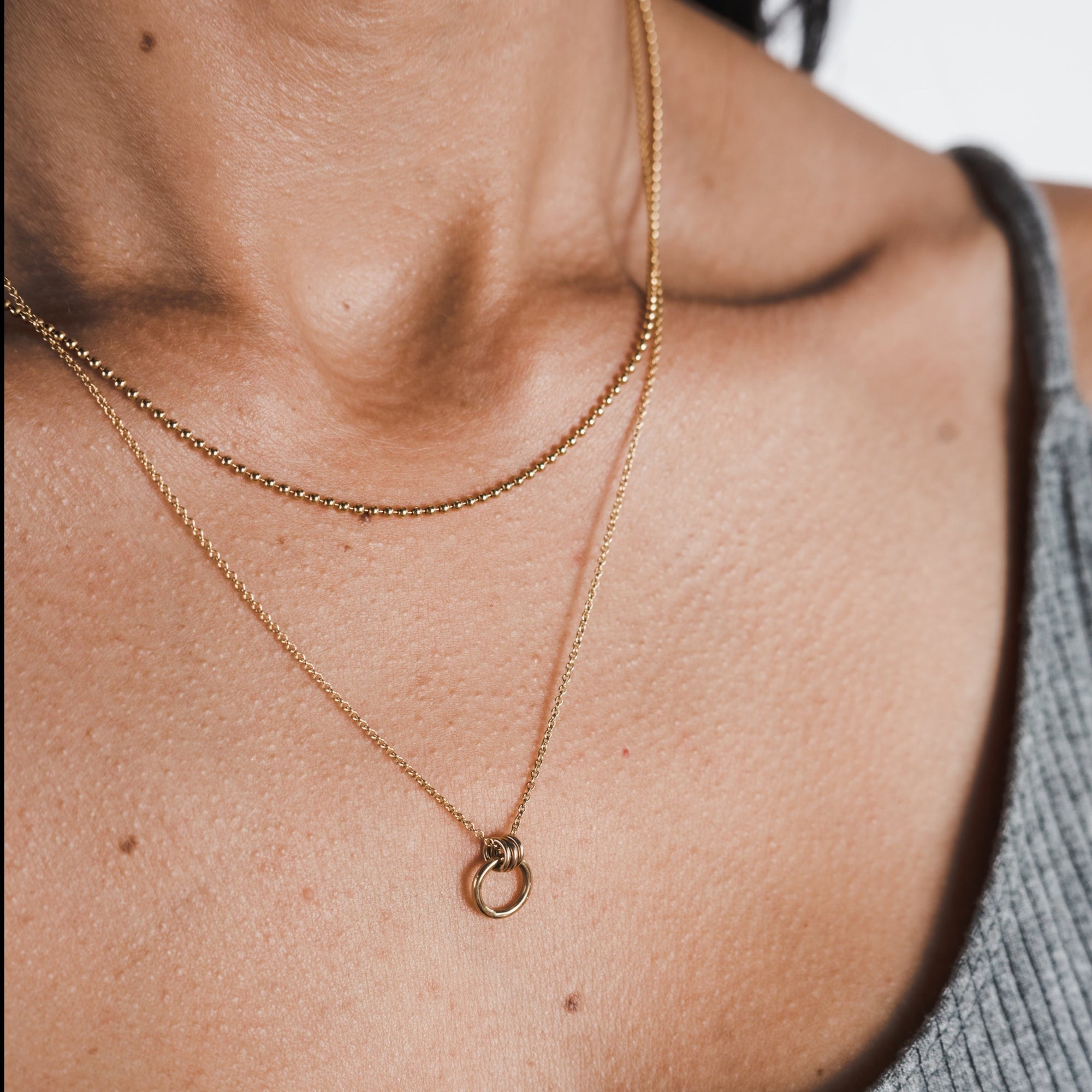 A close-up shot captures a person&#39;s neck beautifully adorned with two delicate, layered gold necklaces from Becoming Jewelry. One is the Bead Chain Necklace, featuring a thin chain that adds a subtle elegance to the look. The other necklace showcases a small circular pendant, adding texture and charm to the ensemble. The person complements the necklaces perfectly with a gray sleeveless top.
