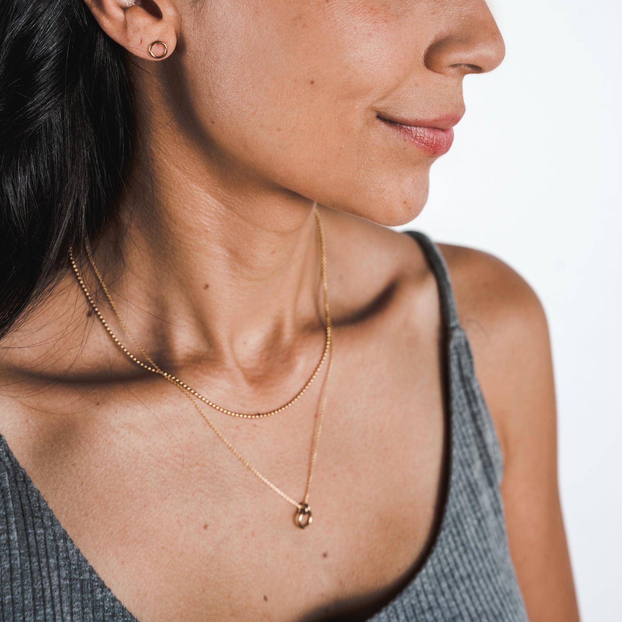 Close-up of a woman wearing a gray top, showcasing gold hoop earrings and two layered necklaces—one is the Bead Chain Necklace from Becoming Jewelry and the other features a small ring pendant, adding texture to her elegant ensemble.