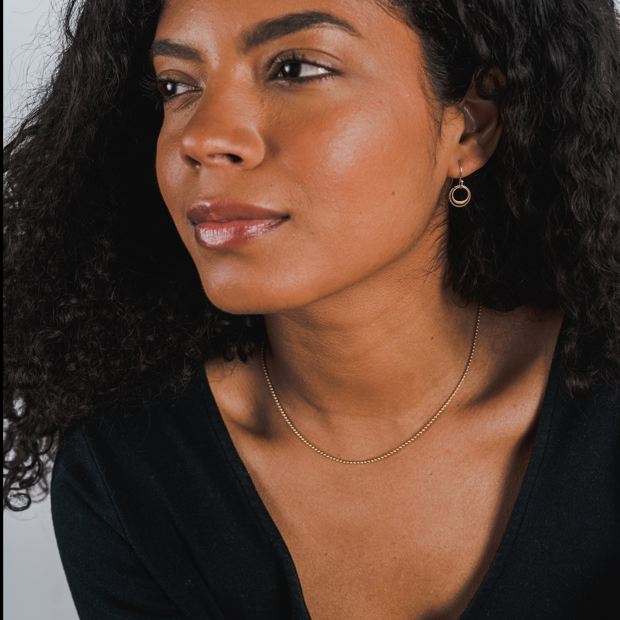 A person with curly hair wearing a black top, a thin gold Bead Chain Necklace from Becoming Jewelry, and a single hoop earring looks to the side against a plain background.